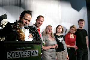 Science-Slam Moderator Christoph Tiemann mit der Trophe, dem "Goldenen Hirn", und den Teilnehmern der vergangenen Veranstaltung.