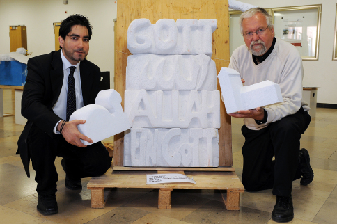 Prof. Dr. Mouhanad Khorchide (l.) und Projektleiter Georg Bienemann vor einem Kunstwerk mit der Aufschrift "Gott - Allah - Ein Gott"