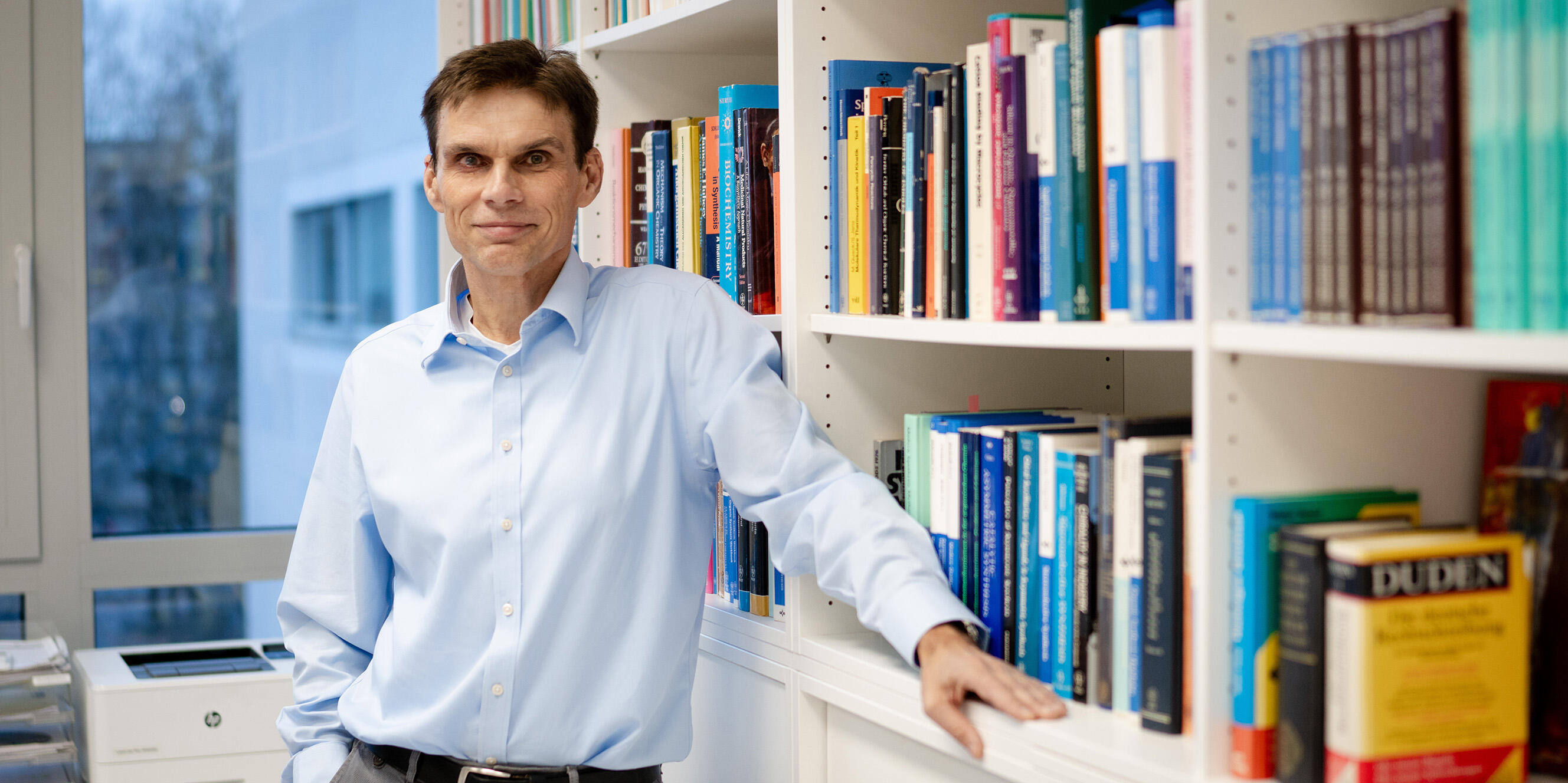Professor Armido Studer lehnt an einem Bücherregal mit chemischen Fachbüchern.