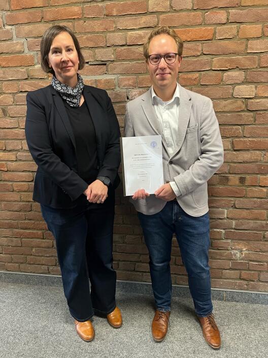 Photo of Nils Stockmann with his doctoral thesis in his hand, and Prof'in Antonia Graf