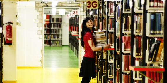 Blick in eine Bibliothek mit Bücherregalen. Eine junge Frau trägt einen Stapel Bücher.