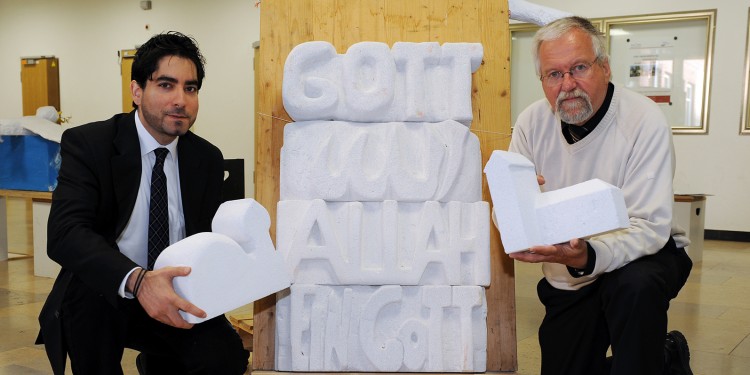 Prof. Dr. Mouhanad Khorchide (l.) und Projektleiter Georg Bienemann vor einem Kunstwerk mit der Aufschrift &quot;Gott - Allah - Ein Gott&quot;<address>© WWU - Peter Grewer</address>