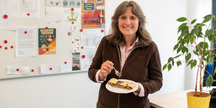 Kerstin Ewers mit einem Stück Mohnkuchen vor einer Pinnwand und einer Grünpflanze in ihrem Büro an der Röntgenstraße.<address>© Uni MS - Johannes Wulf</address>