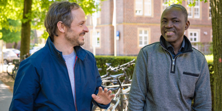 Two men, Professor Frieder Schaumburg (left) and Professor Selidji Todagbe Agnandji, are walking along a street. They are chatting and smiling at each other.<address>© Uni MS - Linus Peikenkamp</address>