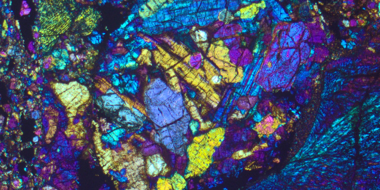 Das Bild zeigt eine mikroskopische Aufnahme eines Dünnschliffs. Die sogenannten Chondren weisen unterschiedliche Farben wie Gelb, Lila, Blau und Grün auf.<address>© Addi Bischoff</address>