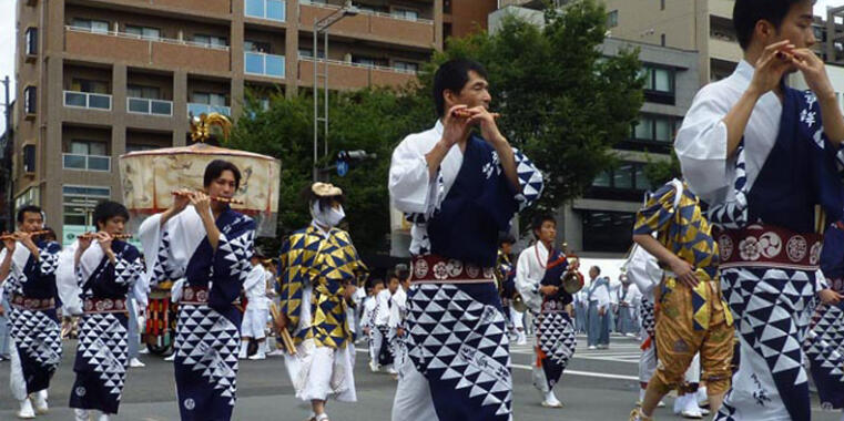A group of people in traditional Japanese attire participating in a festival parade. Several participants are playing traditional flutes. Their robes feature a striking navy blue and white triangle pattern. In the background, modern apartment buildings and a large, decorated festival umbrella are visible.