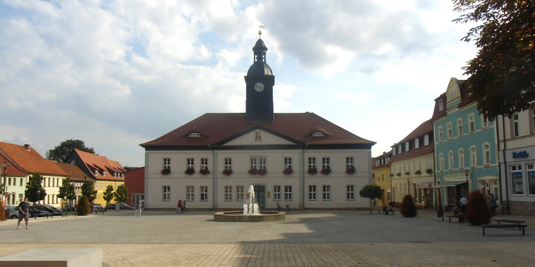 Heller Marktplatz mit Brunnen vor einem historischen Rathaus mit rotem Dach und Uhrturm, umgeben von farbigen Häusern unter einem wolkigen Himmel.