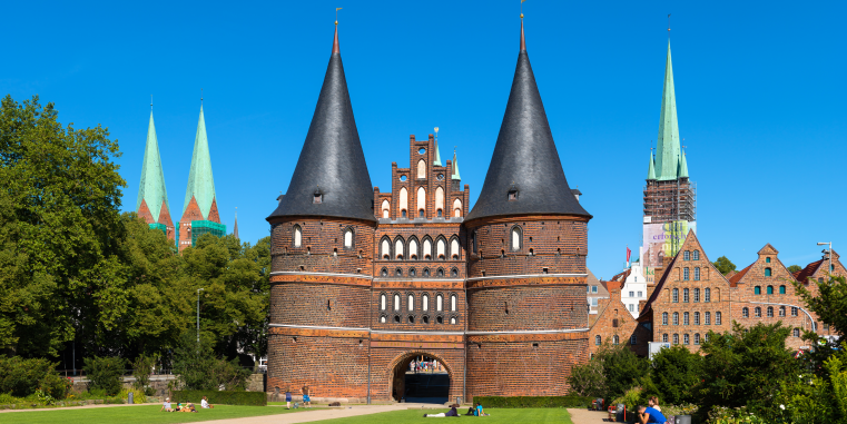 Monumentales mittelalterliches Stadttor aus Backstein mit zwei runden Türmen und spitzen Dächern, davor eine Grünfläche und im Hintergrund mehrere Kirchtürme unter klarem blauem Himmel.