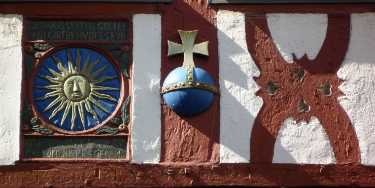 Relief in einer Fachwerkfassade in rot, weiß und blau. Links ist eine goldene Sonne eingelassen.