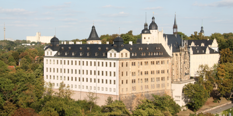 Großes historisches Schloss- oder Klostergebäude mit heller Fassade, dunklen Dächern, Türmen und Kapellenanbau, umgeben von vielen Bäumen unter hellem Himmel.