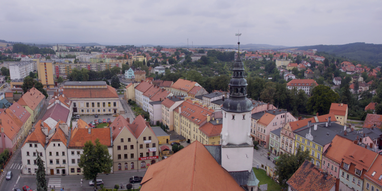 Blick über eine Kleinstadt mit roten Dächern und pastellfarbenen Häusern, dominiert von einem weißen Kirchturm mit dunkler Haube unter bedecktem Himmel.