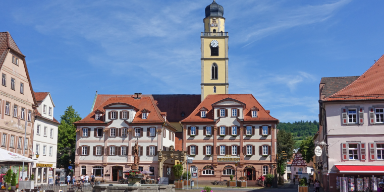 Heller Marktplatz mit Brunnen vor einem historischen Rathaus mit rotem Dach und Uhrturm, umgeben von farbigen Häusern unter einem wolkigen Himmel.
