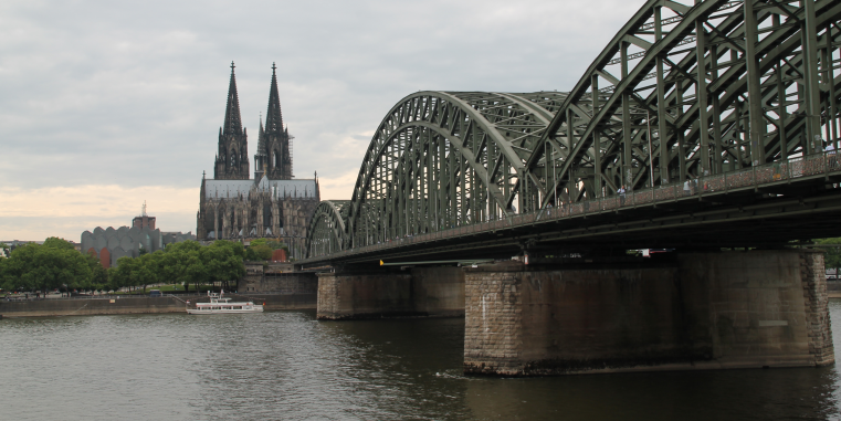 Große Stahlbogenbrücke über einen Fluss mit dem Kölner Dom im Hintergrund unter grauem, bewölktem Himmel.
