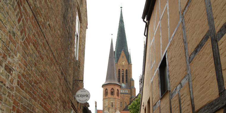 Blick durch eine enge Altstadtgasse zwischen Backstein- und Fachwerkhäusern auf eine hohe Kirche mit spitzem Turm und Uhr.