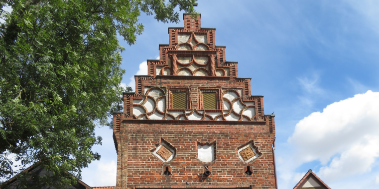 Backsteingiebel eines historischen Gebäudes mit reich gegliederter Staffelgiebel-Form und weißen Schmuckfeldern, eingerahmt von einem Baum und blauem Himmel mit Wolken.