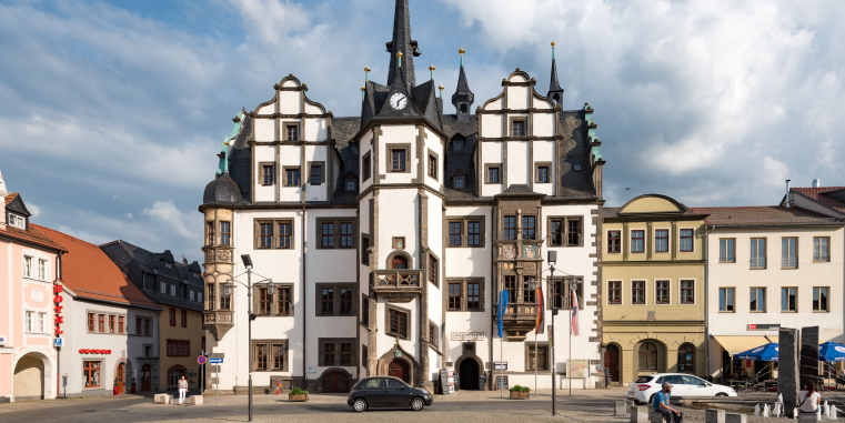 Prunkvolles historisches Rathaus mit heller Fassade, geschwungenen Giebeln, Erkerturm und Uhr auf einem städtischen Platz unter dramatisch bewölktem Himmel.