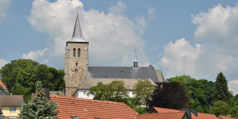 Kirche mit rechteckigem Steinturm und spitzem Dach, leicht erhöht über Häusern mit roten Ziegeldächern und umgeben von Bäumen unter einem wolkigen Himmel.