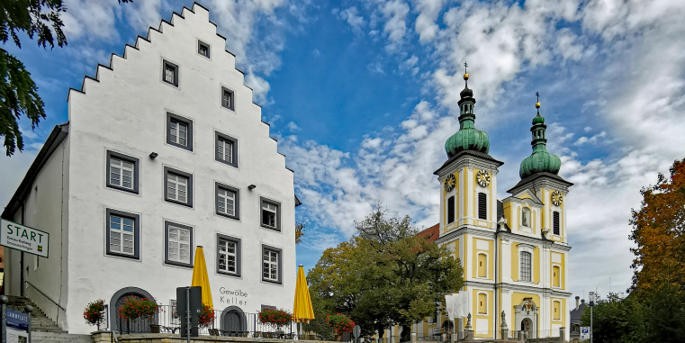 Weißes Giebelhaus und barocke Kirche mit zwei grün gedeckten Zwiebeltürmen an einem Platz unter lebhaft bewölktem blauem Himmel.
