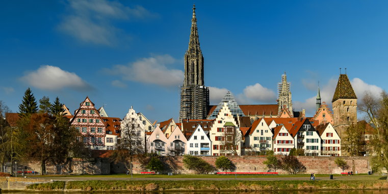 Blick über einen Fluss auf eine historische Stadt mit Giebelhäusern, Stadtmauer, einem gotischen Kirchturm und einem runden Steinturm unter blauem Himmel.