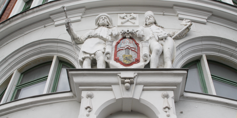 Fassadenschmuck an einem historischen Gebäude mit zwei weißen Bergmannsfiguren, gekreuzten Werkzeugen und einem farbigen Wappen über geschwungenen Fenstern.