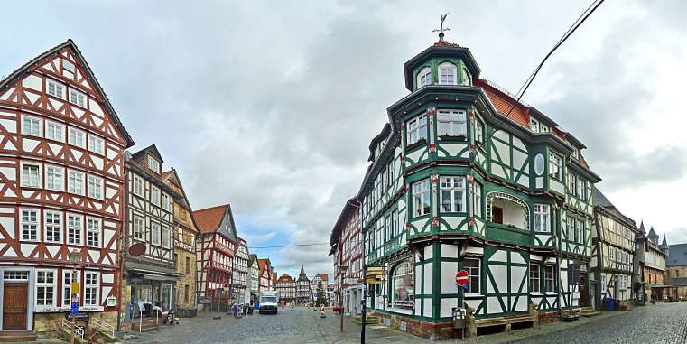 Kopfsteinpflasterstraße in einer Altstadt mit reich verzierten Fachwerkhäusern, darunter ein markantes grün-weißes Eckhaus, unter dramatisch bewölktem Himmel.