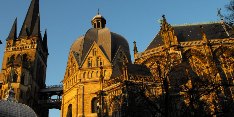 Alttext: Historische Kathedrale mit hohem Kirchturm, großer Kuppel und reich verzierten gotischen Fassaden im warmen Abendlicht vor blauem Himmel; dunkle Baumäste ragen ins Bild.