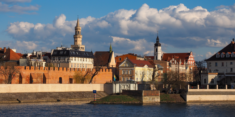 Stadtansicht am Fluss mit Backsteinmauer, historischen Häusern und mehreren Türmen, beleuchtet von warmem Sonnenlicht unter dramatischen Wolken.