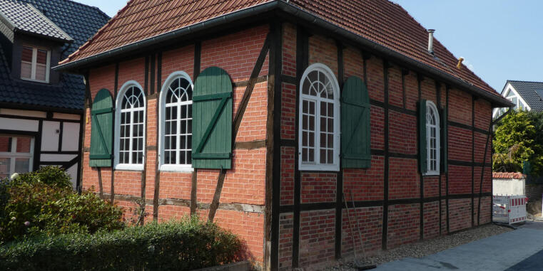 Brick half-timbered building with a red tiled roof, tall arched windows, and green shutters, photographed along a narrow street.