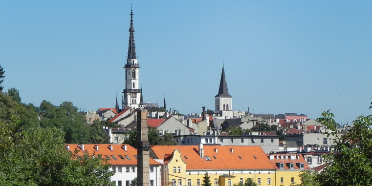 Stadtansicht mit roten Dächern und zwei markanten Kirchtürmen, darunter ein sehr hoher schlanker Turm, über grünen Bäumen unter klarem blauem Himmel.