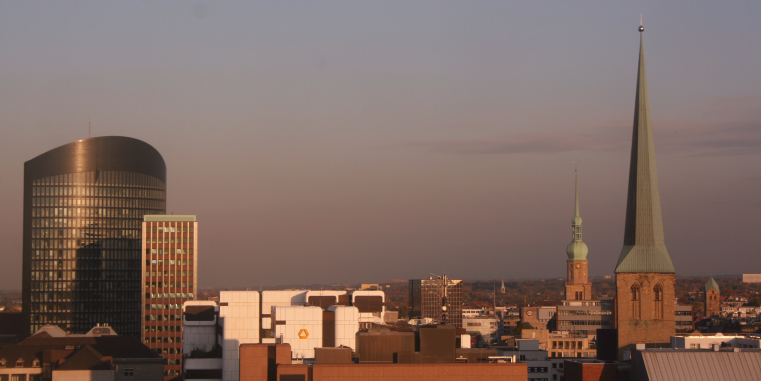 Skyline einer Stadt im warmen Abendlicht mit modernem Hochhaus, Bürogebäuden und zwei markanten Kirchtürmen, darunter ein sehr hoher schlanker Turm.