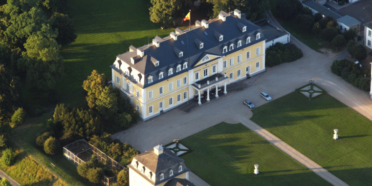 Luftaufnahme eines gelben Schlossgebäudes mit dunklem Dach, Ehrenhof und weitläufigen Rasenflächen, umgeben von Bäumen und Nebengebäuden.