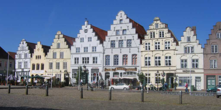 Reihe historischer Giebelhäuser mit hellen Fassaden an einem Marktplatz unter klarem blauem Himmel, davor wenige Menschen und Autos.