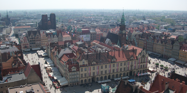 Luftaufnahme einer historischen Altstadt mit großem Marktplatz, Rathaus mit Uhrturm, dicht stehenden Häusern mit roten Dächern und Kirchen im Hintergrund.