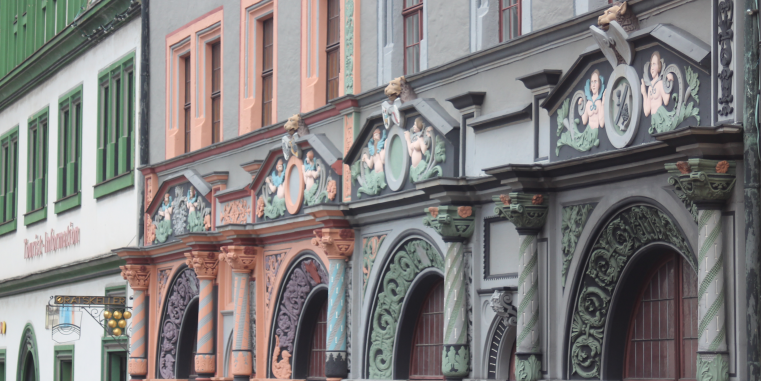 Reich verzierte historische Fassaden mit farbigen Ornamenten, Figuren und Rundbogenportalen entlang einer Häuserzeile.