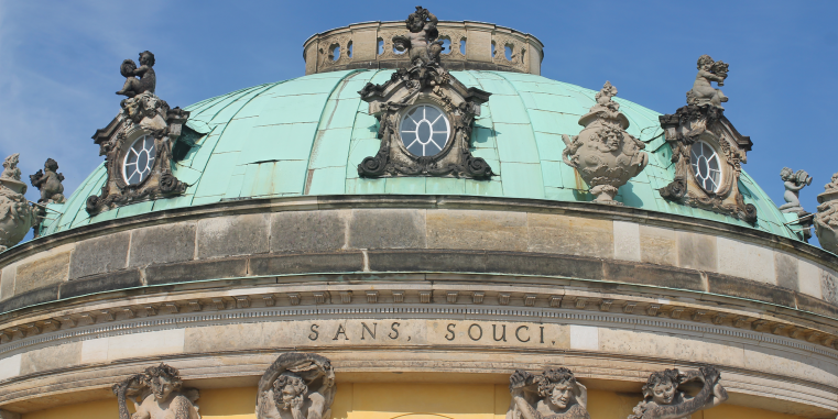 Detailansicht der grünen Kuppel von Schloss Sanssouci mit verzierten Rundfenstern, Puttenfiguren, steinernen Vasen und der Inschrift „Sans, Souci“.
