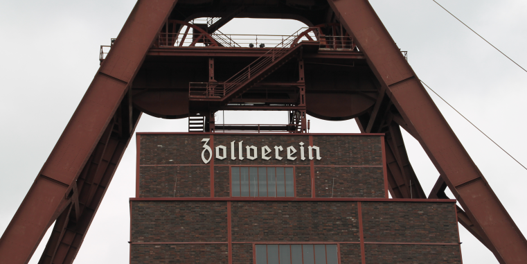 Fördergerüst der Zeche Zollverein aus rostbraunem Stahl über einem Backsteingebäude mit dem Schriftzug „Zollverein“ vor hellem Himmel.