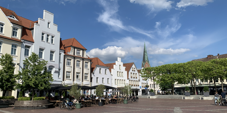 Belebter Marktplatz mit historischen Giebelhäusern, Straßencafés und Bäumen; im Hintergrund ragt ein schlanker Kirchturm in den blauen Himmel mit weißen Wolken.