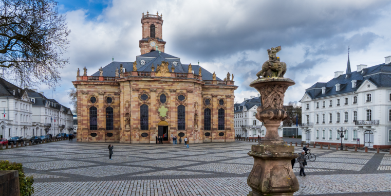 Barocke Kirche aus Sandstein an einem weiten gepflasterten Platz, flankiert von hellen Gebäuden; im Vordergrund steht eine reich verzierte steinerne Brunnen- oder Säulenskulptur.