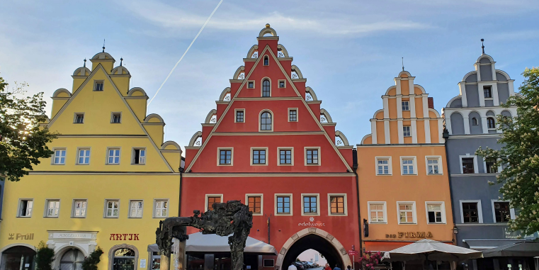 Reihe farbenfroher historischer Giebelhäuser mit geschwungenen Staffelgiebeln an einem Platz, darunter Läden und Cafés, vor blauem Himmel.