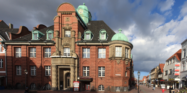 Historisches rotes Backsteingebäude mit aufwendiger Fassade, grünen Kupferkuppeln und Uhrturm an einer Einkaufsstraße unter dramatischem Wolkenhimmel.
