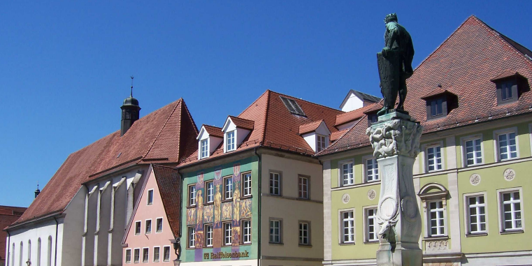 Historischer Platz mit farbigen Häuserfassaden, einer Kirche mit steilem Dach und einer Statue auf hoher Säule vor klarem blauem Himmel.