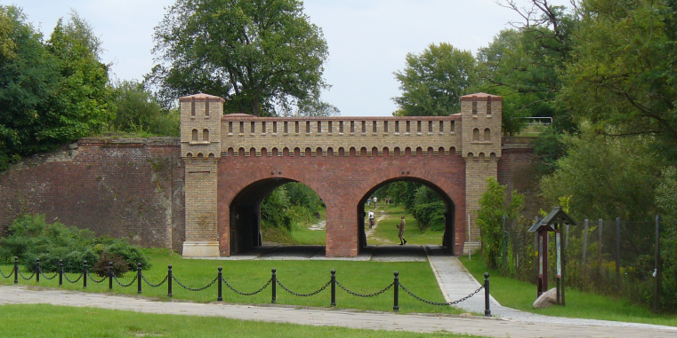 Historisches Backsteinbauwerk mit zwei Rundbogen-Durchgängen und zinnenartigem Aufbau, eingebettet in eine grüne Park- oder Wallanlage mit Bäumen und Wegen.