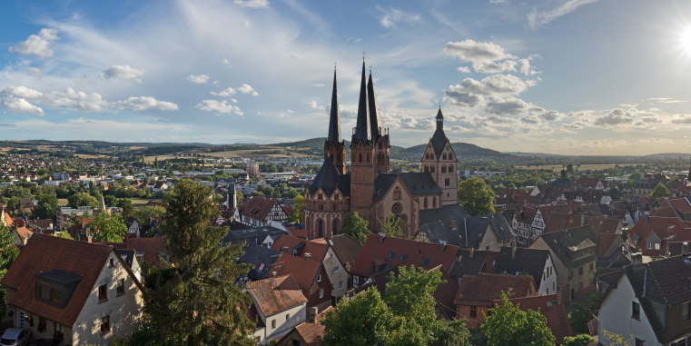Panorama einer Kleinstadt mit roten Dächern und großer Kirche mit drei hohen Türmen, umgeben von hügeliger Landschaft unter sonnigem Himmel mit vielen Wolken.