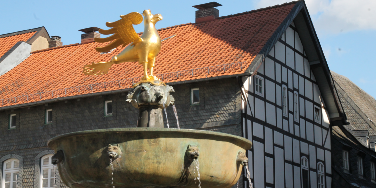 Brunnen mit goldener Vogelstatue vor historischen Fachwerkhäusern mit dunklem Holz und roten Ziegeldächern im Sonnenlicht.
