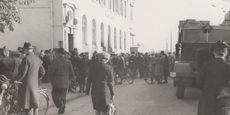 Blick in eine Straßen, auf der linken Seite befindet sich ein großes, weißes Gebäude. Davor befinden sich zahlreiche Menschen in Mänteln, die Mäner und einige der Frauen tragen Hüte, einige der Personen haben ein Fahrread. Auf der rechten Seite befindet sich ein großes Fahrzeug, das darauf hinweist, dass das Foto aus den 1930er bis 1960er Jahren kommt. Das Foto ist schwarz-weiß.