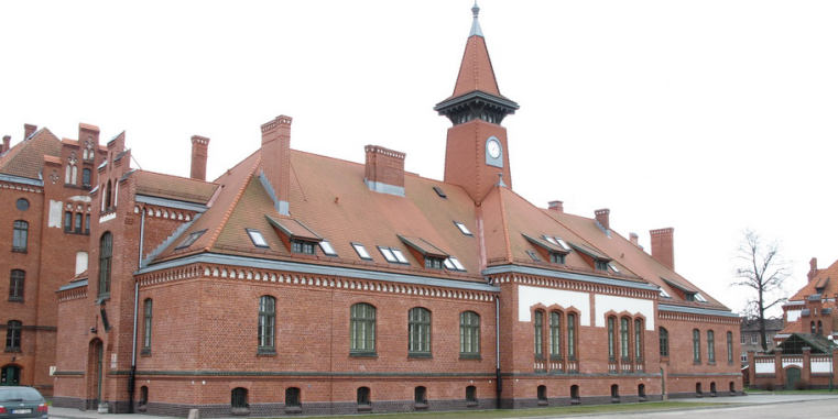Historisches Backsteingebäude mit langgezogenem Satteldach, vielen Schornsteinen und einem kleinen Uhrturm, aufgenommen von der Straßenseite.