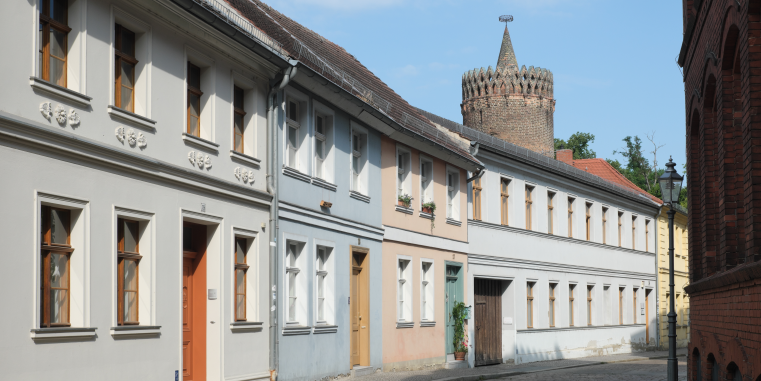 Ruhige Altstadtgasse mit hellen historischen Häusern, schmalen Fenstern und pastellfarbenen Türen; dahinter ragt ein runder Backsteinturm mit gezackter Krone in den blauen Himmel.