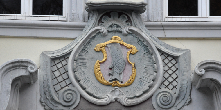 Nahaufnahme eines kunstvollen Fassadenschmucks an einem Gebäude: In der Mitte befindet sich ein Wappenschild mit einem Fisch, umgeben von ornamentalen Verzierungen in Grau und Gold.