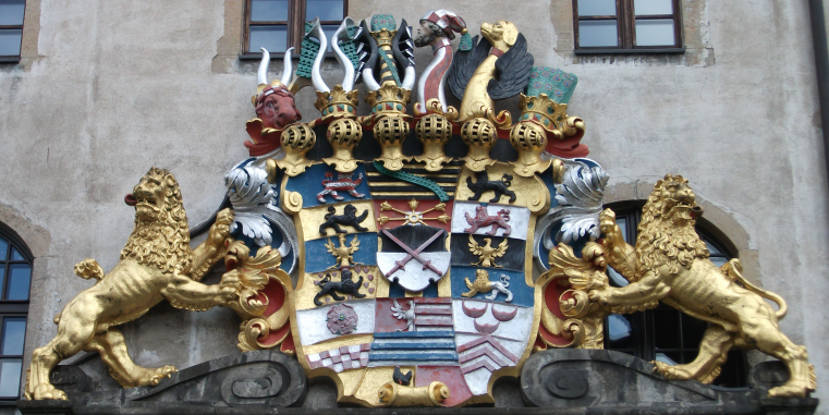 Prunkvolles farbiges Wappenrelief an einer historischen Fassade, flankiert von zwei goldenen Löwen und bekrönt von mehreren Helmzierden.
