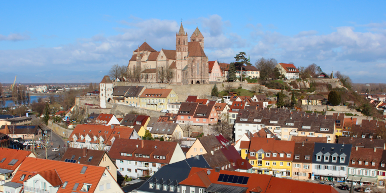 Blick über eine Stadt mit roten Dächern zu einer erhöht gelegenen romanischen Kirche und befestigten Anlage auf einem Hügel, unter blauem Himmel mit Wolken.